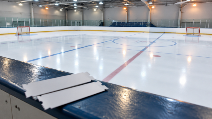 Blick von der Tribüne einer kleinen, voll beleuchteten Eishalle auf eine leere Eisfläche mit roten Toren, davor Ticketabschnitte auf der Bande